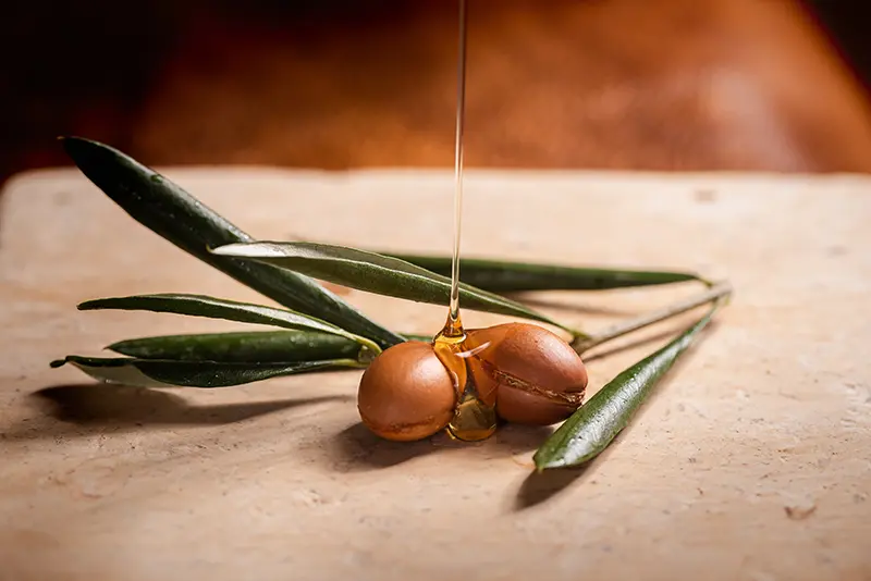 Noci di Argan mature con foglie verdi e olio d'oro che viene versato sopra, simboleggiando la purezza dell'estrazione Kipi.